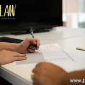 Person signing documents at a white desk in a law office, with a large monitor in the background and the JZW LAW logo visible in the top-left corner.