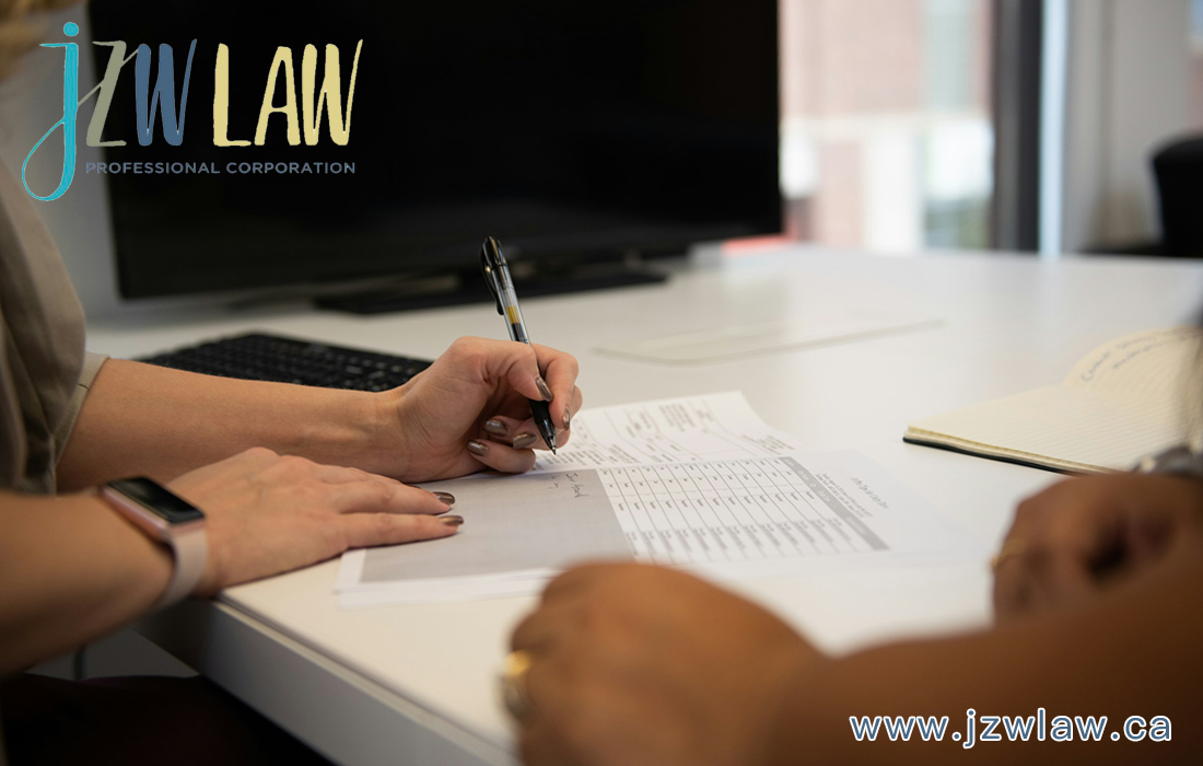 Person signing documents at a white desk in a law office, with a large monitor in the background and the JZW LAW logo visible in the top-left corner.