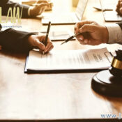 Lawyers signing a contract at a wooden table with a gavel in the foreground; logo and website visible on the image.