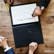Top-down view of a contract being stamped on a wooden desk, with two people clasping hands nearby and rings inside an open folder (law firm branding visible).