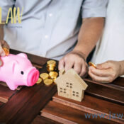 Pink piggy bank with coins and a small wooden house model on a dark wooden table; two hands placing coins, with JZW Law branding and the site www.jzwlaw.ca in view.
