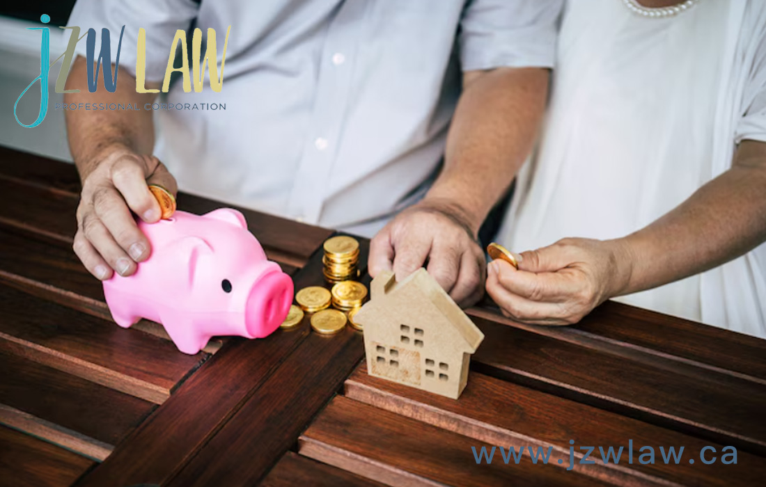 Pink piggy bank with coins and a small wooden house model on a dark wooden table; two hands placing coins, with JZW Law branding and the site www.jzwlaw.ca in view.