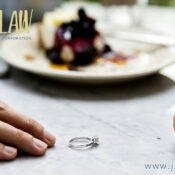 Engagement ring rests on a marble tabletop between two hands, with a blurred dessert plate in the background; law firm branding in the top-left corner and website text in the bottom-right.