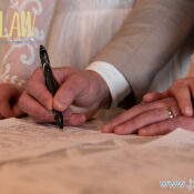 Close-up of a couple signing documents together, hands on the paper with wedding rings visible and a pen between them, logo in the top-left corner.