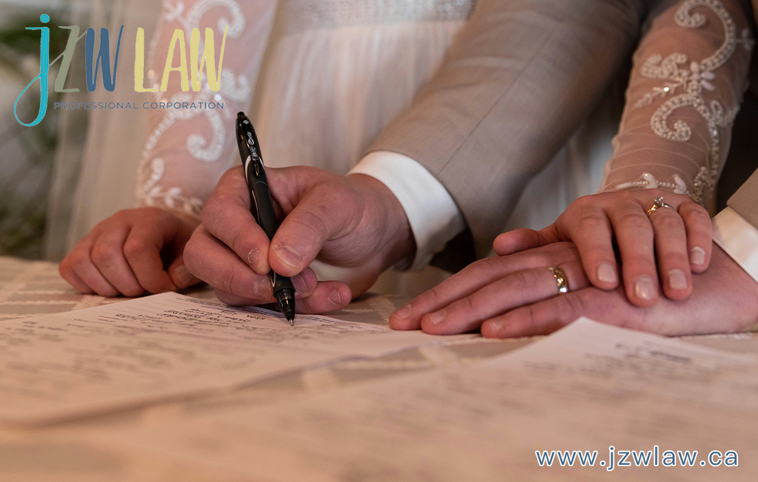 Close-up of a couple signing documents together, hands on the paper with wedding rings visible and a pen between them, logo in the top-left corner.