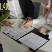 Two professionals sign and discuss documents at a white table in a law office, with a pen over a contract open in a binder.