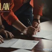 Hands signing documents on a wooden table, law firm branding visible in the corner.